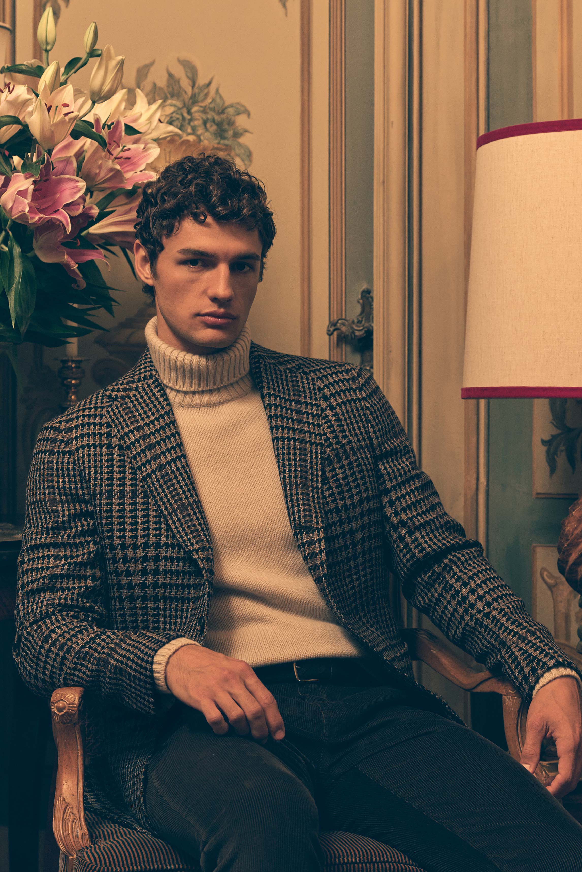 Front view of a seated male model wearing taupe and black checkered blazer 'Vincenzo' by Stile Latino, sand cashmere turtleneck by Bruno Manetti, dark brown corduroy 'Bobby' pants from Jacob Cohen, and dark brown braided belt by Officine Creative. The Sho