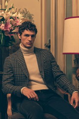 Front view of a seated male model wearing taupe and black checkered blazer 'Vincenzo' by Stile Latino, sand cashmere turtleneck by Bruno Manetti, dark brown corduroy 'Bobby' pants from Jacob Cohen, and dark brown braided belt by Officine Creative. The Sho