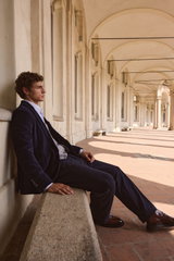 Side view of a model seated on a stone bench in an arched corridor, dressed in a navy velvet blazer and pants from Circolo 1901, grey Malo cashmere cardigan, light blue striped 'Nello' shirt by Fray, and brown 'Dude Flexi' desert boots by Officine Creativ