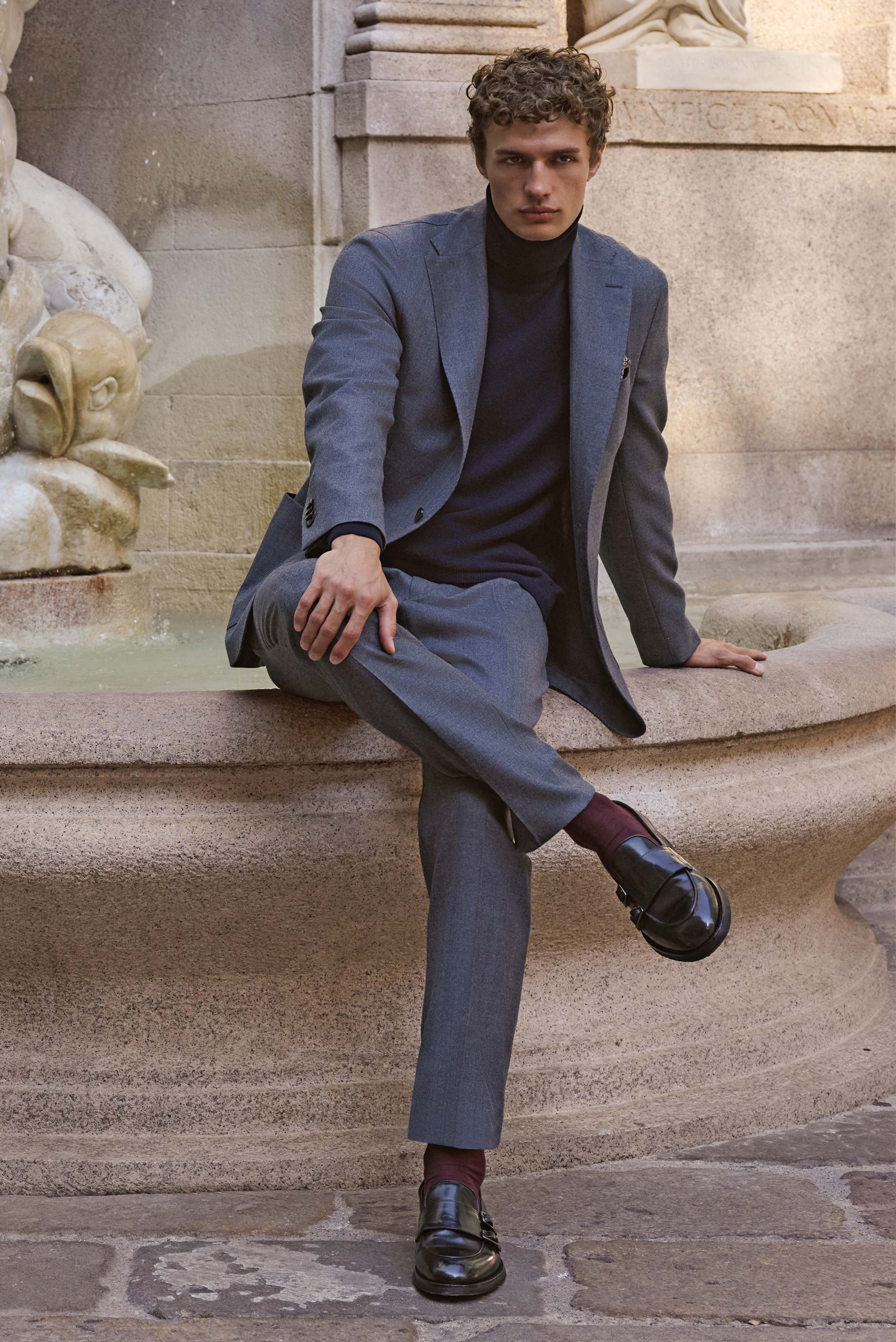 Front view of a seated male model wearing a navy cashmere turtleneck sweater from Aida Barni, paired with a grey suit and black Double Monk 'Opera Flexi' shoes from Officine Creative, accessorized with a mauve and dark blue patterned silk pocket square fr