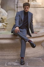 Front view of a seated male model wearing a navy cashmere turtleneck sweater from Aida Barni, paired with a grey suit and black Double Monk 'Opera Flexi' shoes from Officine Creative, accessorized with a mauve and dark blue patterned silk pocket square fr