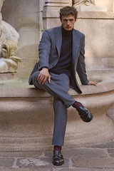 Front view of a seated male model wearing a navy cashmere turtleneck sweater from Aida Barni, paired with a grey suit and black Double Monk 'Opera Flexi' shoes from Officine Creative, accessorized with a mauve and dark blue patterned silk pocket square fr
