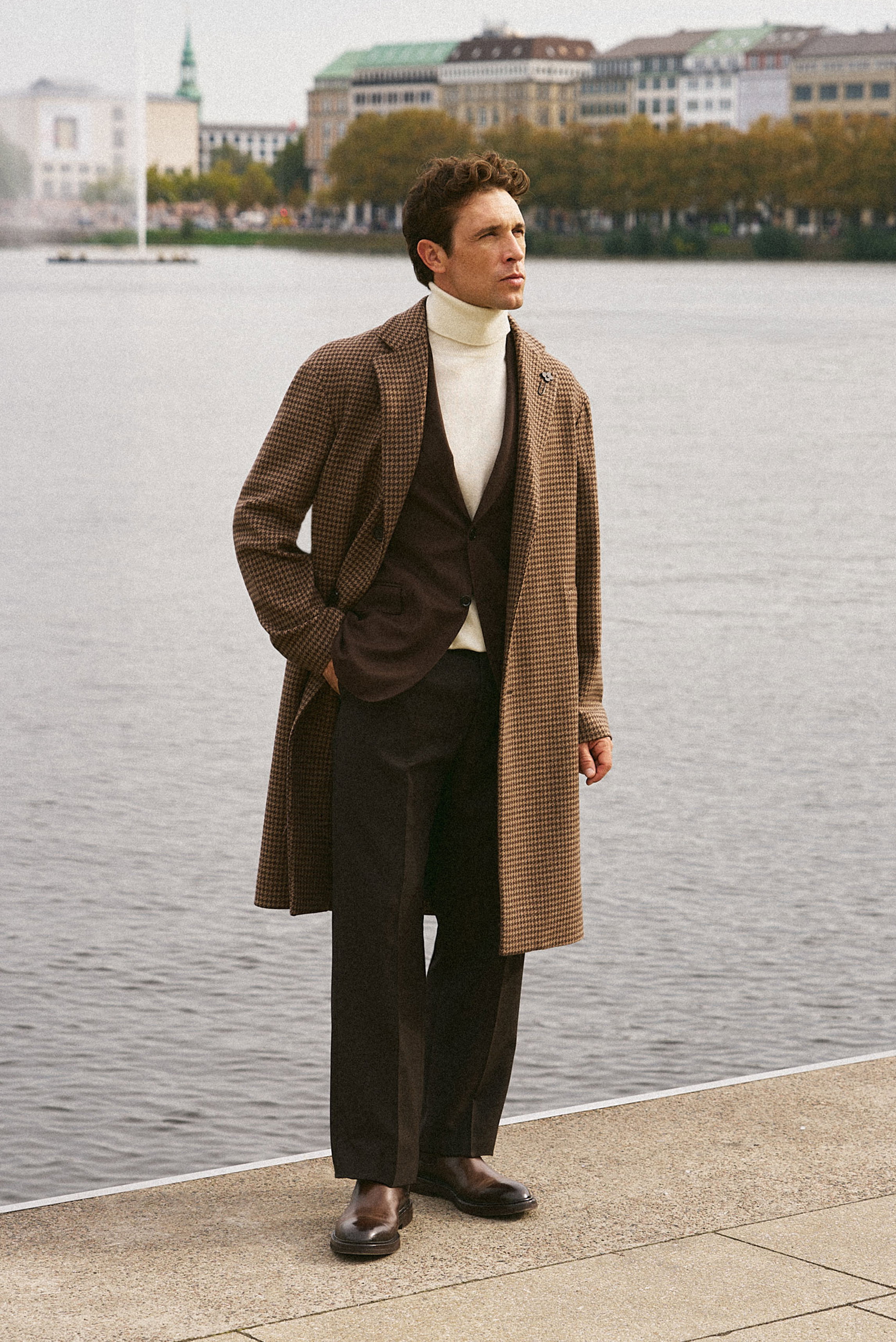 Shop by Look 04 Denis Cebulec in Lardini for BRAUN Hamburg, seitliche Nahaufnahme des Modells mit braun/beigem Mantel, braunem Sakko, creme Rollkragen, Wollhose und braunen Chelsea Boots.