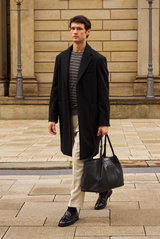 Shop by Look 01 KW 30 2026, front view of a man wearing a black Barena Venezia coat over a navy-grey Emporio Armani sweater, sand PT Torino trousers, black Officine Creative tassel loafers, and carrying a black Serapian shopper bag.