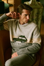 Man seated in a chair seen from a front-side angle wearing a light grey, beige, and grey patterned crewneck sweater from Jacob Cohen's 'Ski' collection. The model is wearing a cozy, soft textured sweater featuring a green car carrying a Christmas tree on 