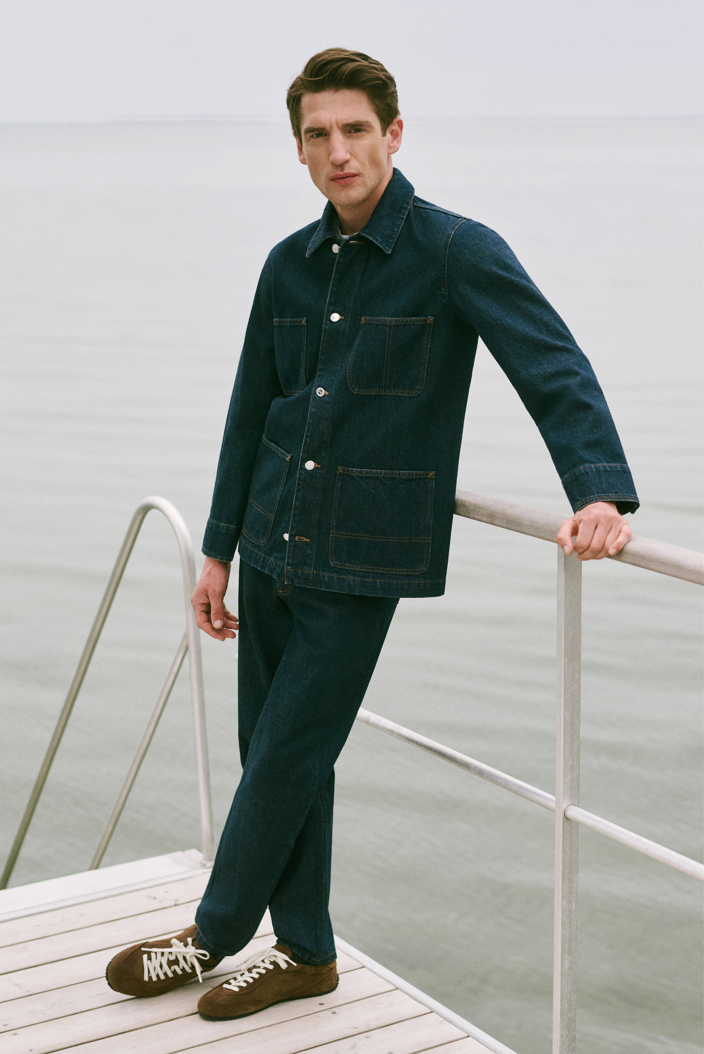 Mann lehnt am Geländer, trägt dunkelblaue Jeansjacke von A.P.C. mit vier Taschen, dunkle Hose, braune Sneaker, Shop by Look 16 HW 2026, Frontansicht.