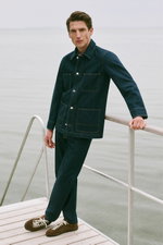 Shop by Look 16 HW 2026: Model photographed from front leaning on railing, wearing navy denim jacket from A.P.C. with classic collar, button-up front, four large patch pockets, dark wash stitching, matching dark denim pants, and brown suede sneakers with 