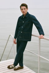 Shop by Look 16 HW 2026: Model photographed from front leaning on railing, wearing navy denim jacket from A.P.C. with classic collar, button-up front, four large patch pockets, dark wash stitching, matching dark denim pants, and brown suede sneakers with 