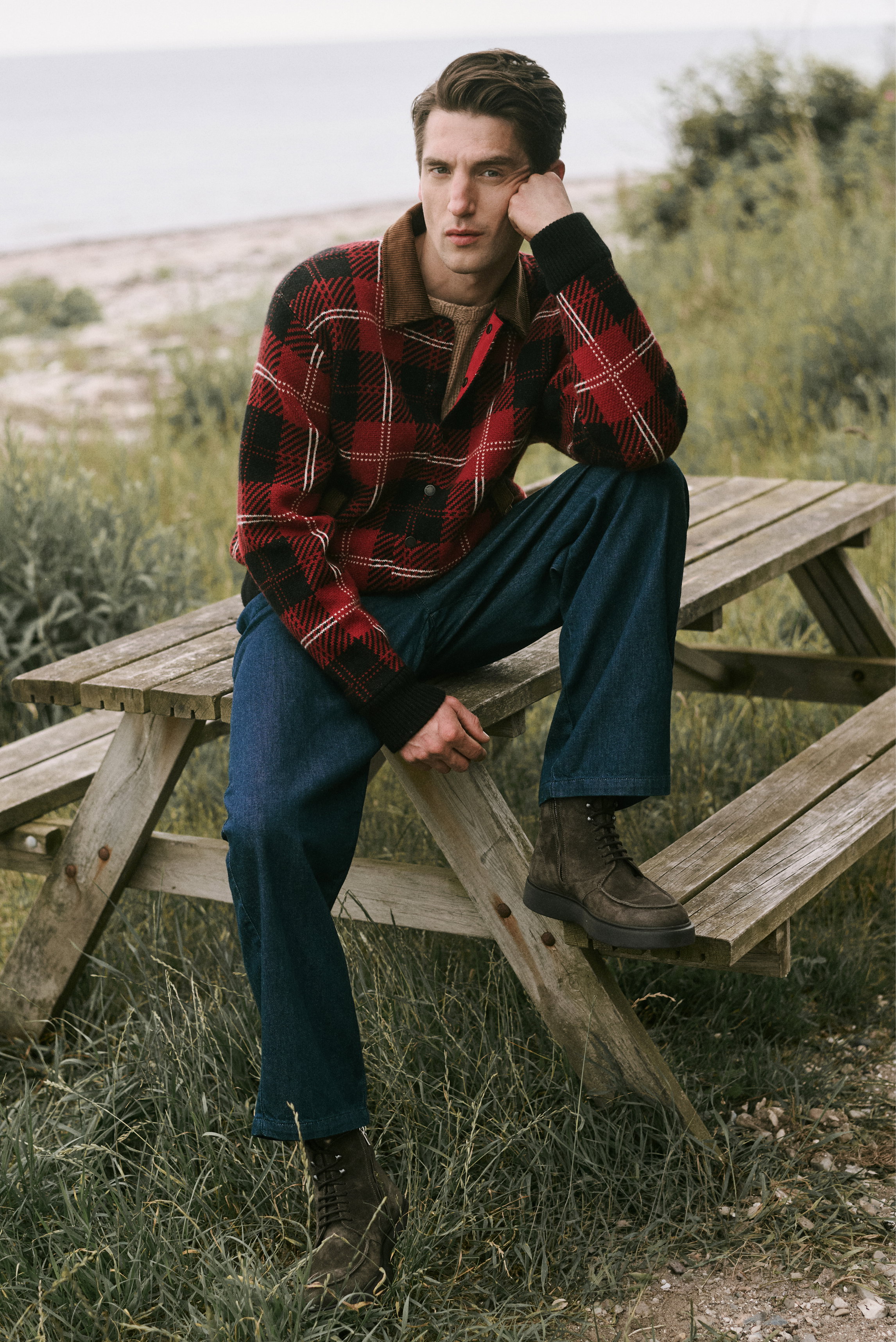 Männliches Model sitzt auf Holztisch, trägt dunkelblaue Baumwollhose „Ricky“ von A.P.C., dunkelbraune Schnürboots „Frame“ von Officine Creative, rotschwarzen karierten Pullover – Shop by Look 17 HW 2026, frontale Perspektive.