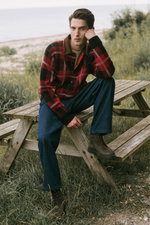 Man sitting on a wooden picnic bench outdoors, photographed from the front-left. The model wears dark blue cotton pants named 'Ricky' from A.P.C., dark brown lace-up 'Frame' boots from Officine Creative, a red and black checkered jacket with brown collar 