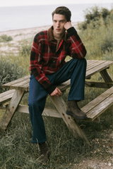 Man sitting on a wooden picnic bench outdoors, photographed from the front-left. The model wears dark blue cotton pants named 'Ricky' from A.P.C., dark brown lace-up 'Frame' boots from Officine Creative, a red and black checkered jacket with brown collar 