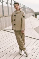 Mann frontal stehend, trägt beige Dufflecoat-Jacke mit Kapuze, schwarzen Knebelverschlüssen, Cargo-Hose mit Taschen, weiße Sneaker, Shop by Look 20 HW 2026.