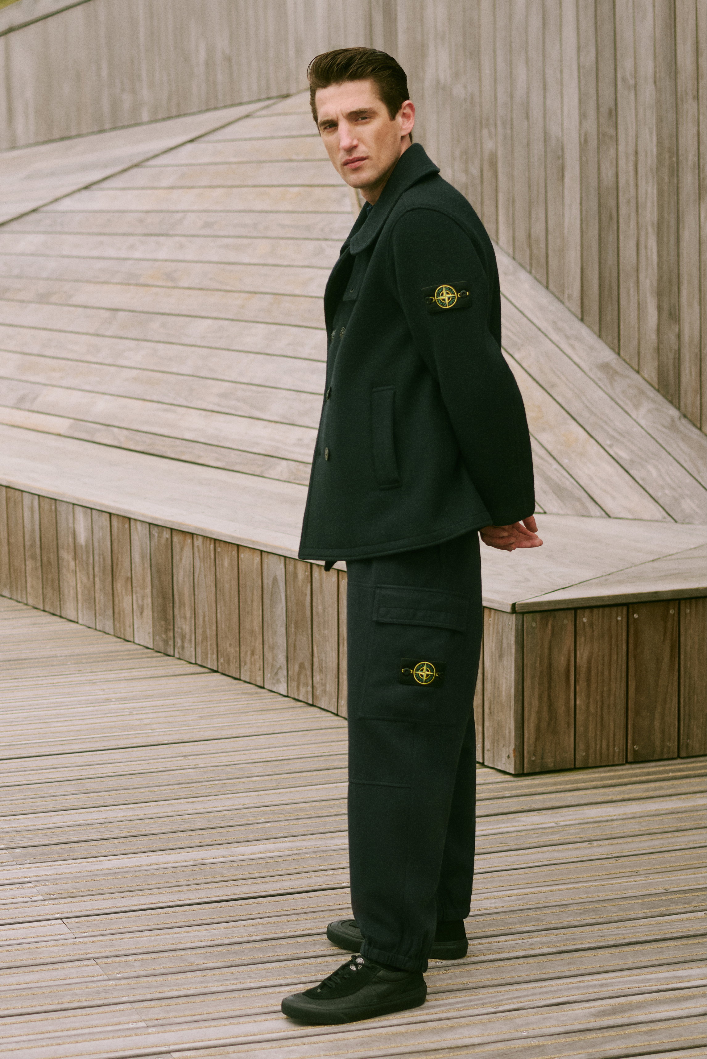 Mann in Dreiviertelansicht trägt schwarzen Mantel mit gelb-grünem Stone Island Patch am Ärmel, schwarze Hose mit Taschen und Sneaker – Shop by Look 21 HW 2026.