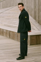 Mann in Dreiviertelansicht trägt schwarzen Mantel mit gelb-grünem Stone Island Patch am Ärmel, schwarze Hose mit Taschen und Sneaker – Shop by Look 21 HW 2026.