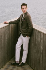 Mann lehnt seitlich an Holzwand mit Blick aufs Wasser, trägt beigen Cashmere Mockneck, cremefarbene Chino, karierten Blazer mit Einstecktuch – Shop by Look 22 HW 2026.