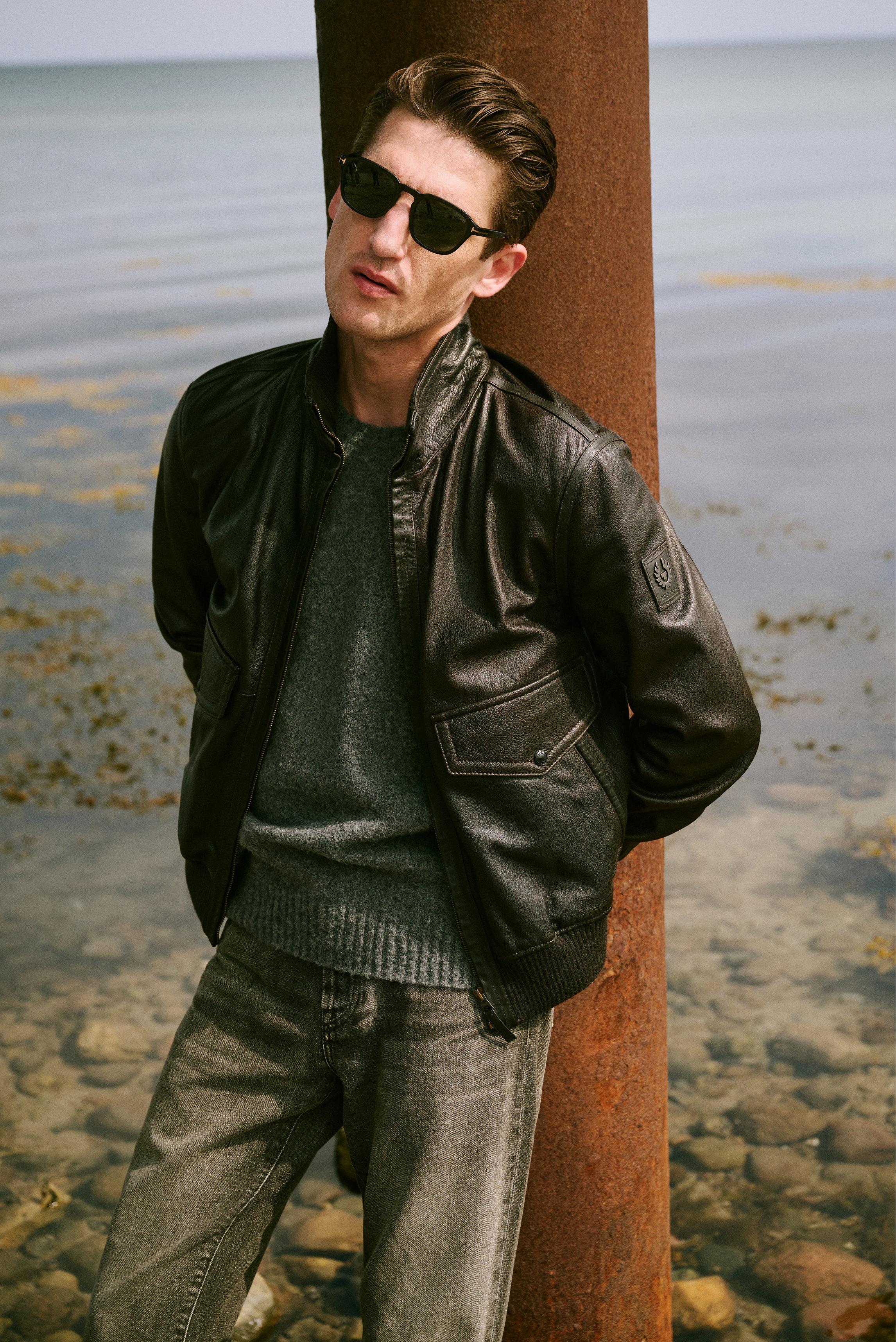 Shop by Look 25 HW 2026 from front angle, model wearing black leather jacket with sleeve patch, grey knit sweater, black sunglasses, and grey washed jeans. Casual yet stylish for cooler weather.