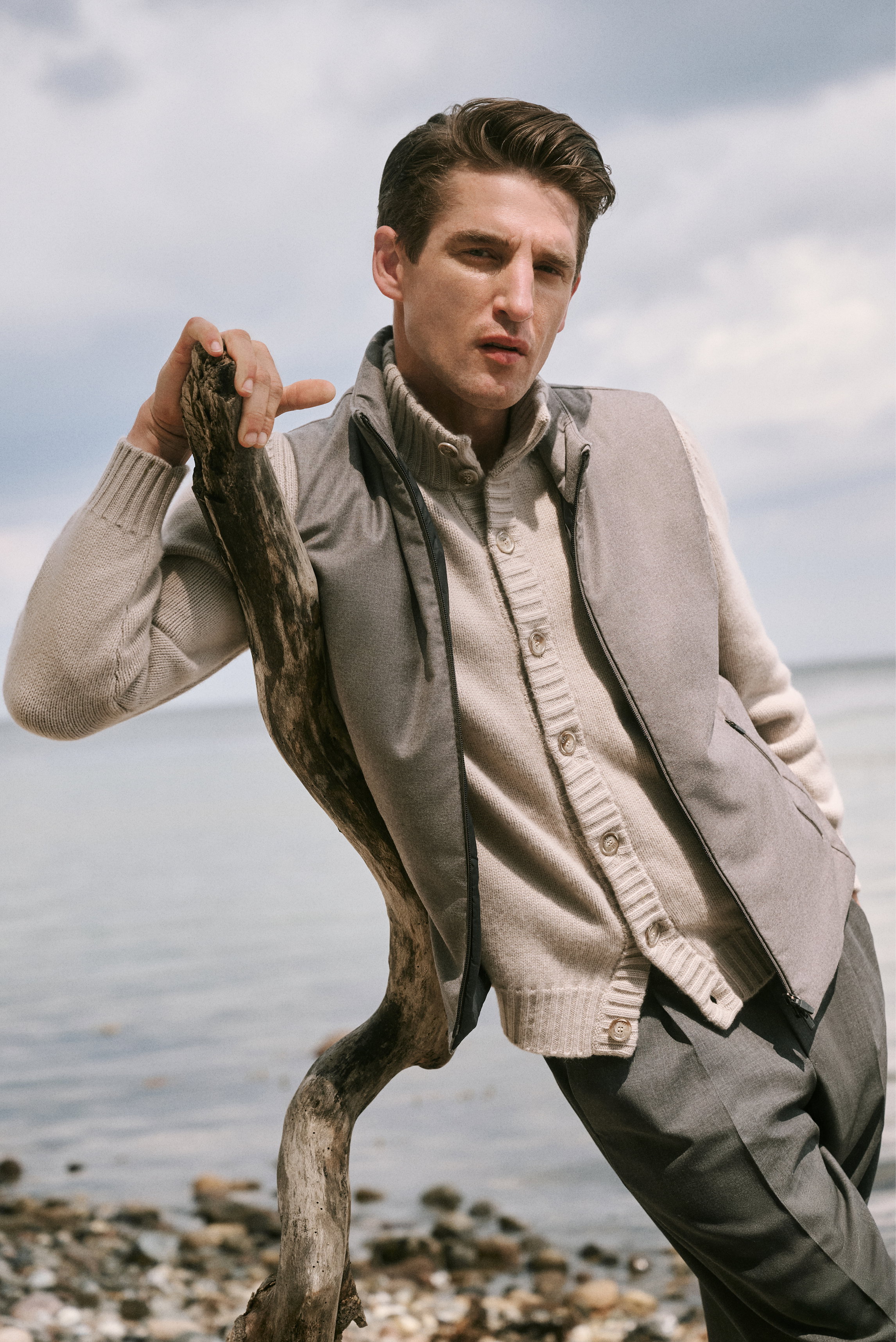 Front view of a man leaning on driftwood by the shore, wearing Shop by Look 28 HW 2026: taupe cashmere 'Favonius' cardigan by Fedeli under a light grey vest, paired with dark grey 'Rebel Fit' wool pants from PT Torino, combining comfort with sophisticated