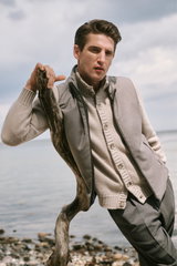 Mann lehnt am Strand an Treibholz, trägt taupefarbenen Kaschmir-Cardigan „Favonius“ von Fedeli und dunkelgrau melierte Wollhose „Rebel Fit“ von PT Torino, seitliche Perspektive, stilvoller Look „Shop by Look 28 HW 2026“.