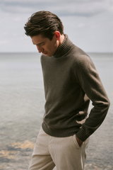 Männliches Modell seitlich fotografiert am Meer trägt graubraunen Cashmere-Rollkragenpullover „Duvet“ von Fioroni und sandfarbene Cordhose „Davide“ von GTA, Shop by Look 29 HW 2026.