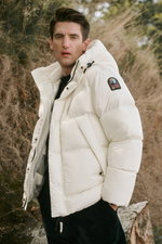 Porträtaufnahme von vorne seitlich des Models mit weißer wattierter Jacke mit Kapuze, navyblauem Rundhalspullover „Edmond“ von A.P.C., navyblauer Cordhose „Rebel Fit“ von PT Torino und dunkelbraunen Schnürboots „Frame“ von Officine Creative, Teil des „Sho