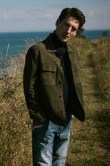 Porträtaufnahme aus schräger Front eines Mannes in Shop by Look 37 HW 2026 mit blauer Jeans 'Bray' von Dondup, schwarzem Rollkragenpullover und olivgrüner, wildlederartiger Jacke mit Brusttaschen, im Freien an Küste.