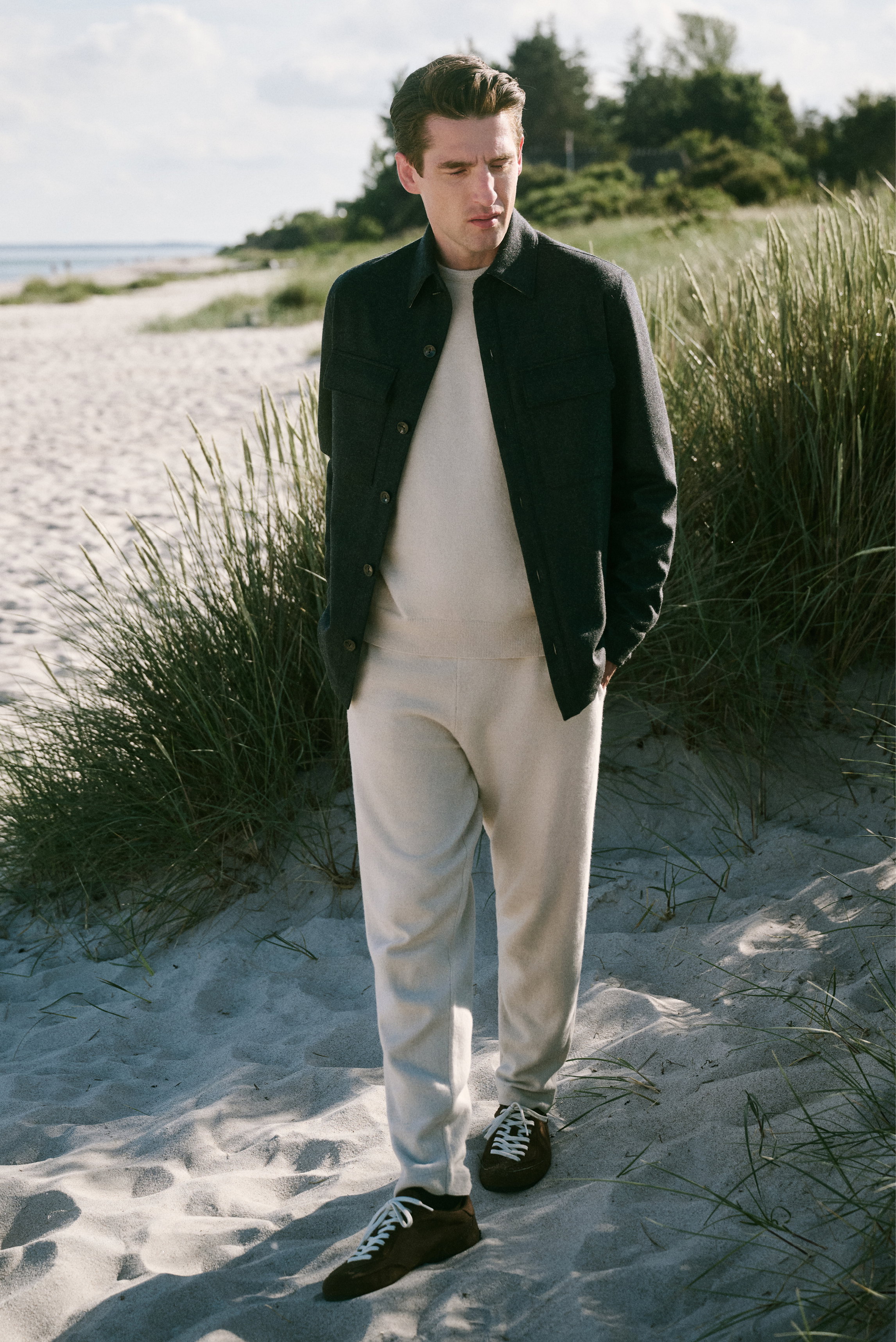 Mann im Profil am Strand mit dunkelgrauer Jacke, hellbeigem Pullover und Hose sowie dunkelbraunen Schuhen, Shop by Look 39 HW 2026.