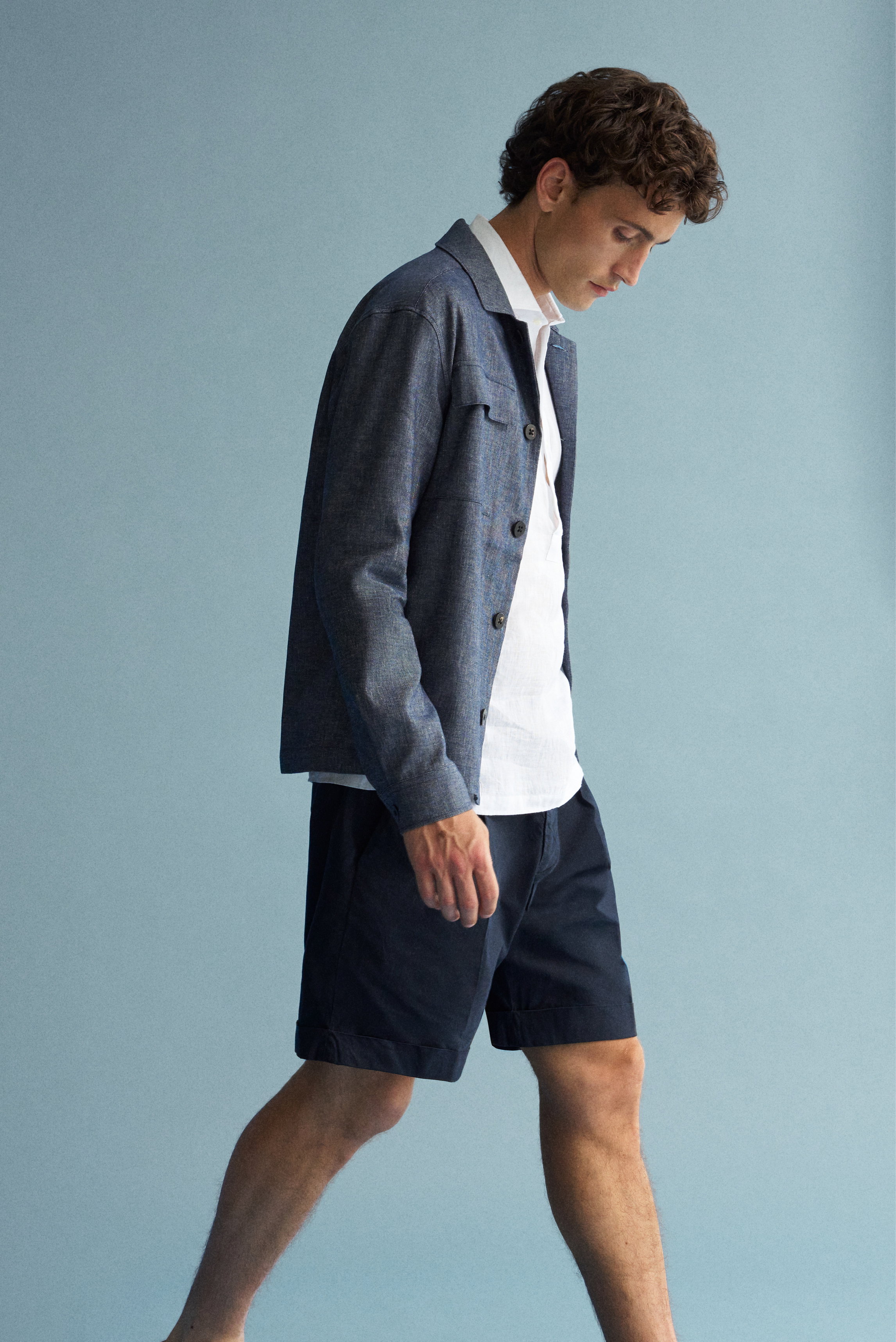 Seitliche Perspektive eines Models in navy meliertem Shop by Look 02 Overshirt „Denim“ von 04651/A trip in a bag, weißem Leinen Popover-Hemd von Giampaolo, navy Shorts „Ethan“ von Dondup und graubraunen „Herbie“ Slippern von Officine Creative, lässig und 