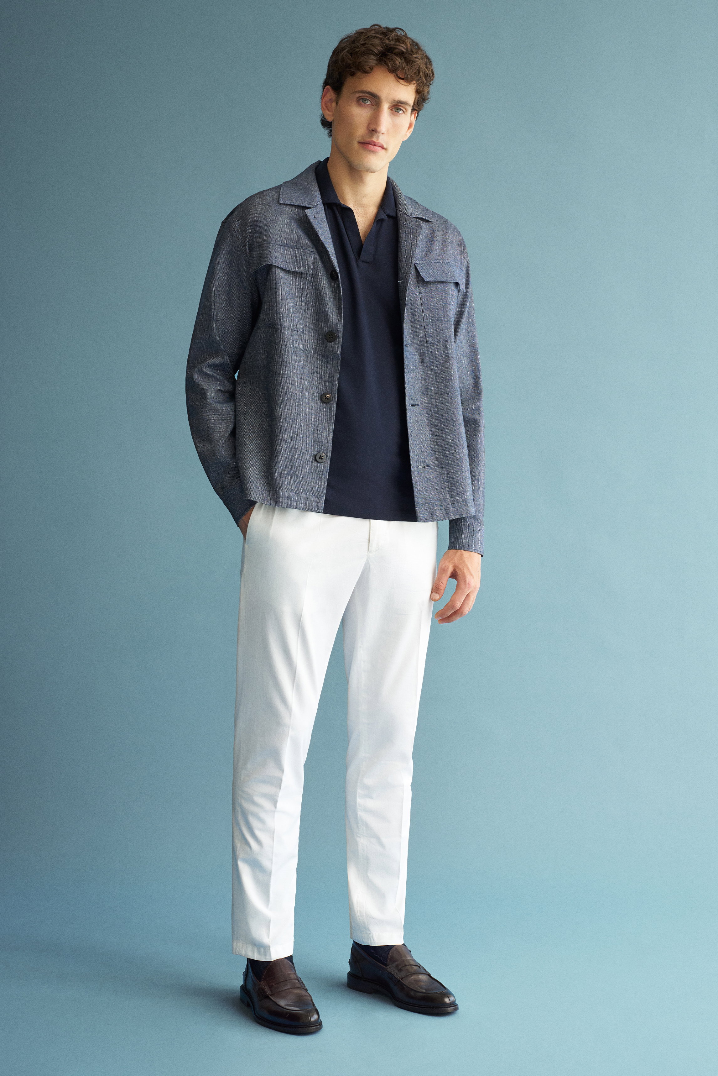 Mann frontal stehend in navy meliertem Shop by Look 01 Overshirt „Denim“ 04651/ A trip in a bag, navy Jersey-Poloshirt, offwhite Chino und dunkelbraunen Loafern, moderner eleganter Casual-Look.