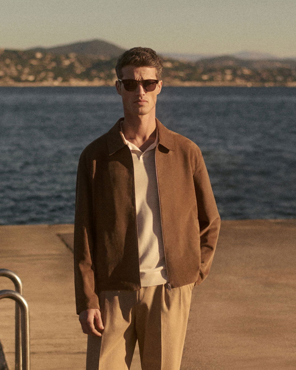Mann mit brauner Jacke, weißem Poloshirt und beiger Hose steht mit Sonnenbrille am Wasser, Berge im Hintergrund.