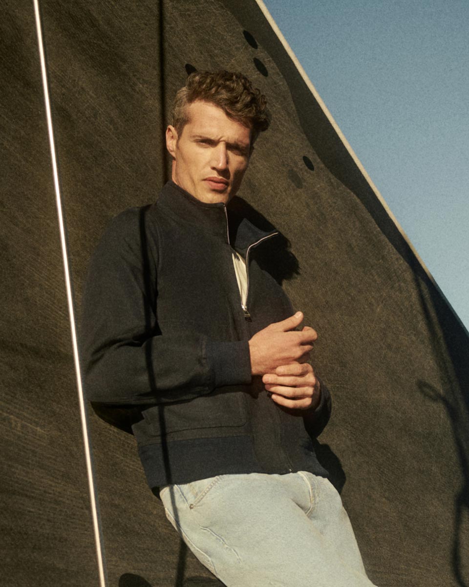Young man with short curly hair wearing a black zip-up jacket and light blue jeans, leaning against a dark textured sail.