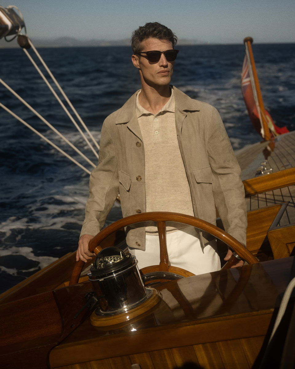 Man wearing sunglasses and beige jacket steering a wooden sailboat on a sunny day with deep blue sea in the background.