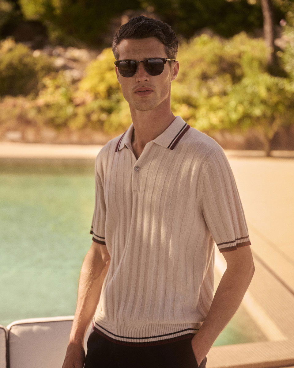 Junger Mann mit dunklen Haaren und Sonnenbrille trägt beiges, kurzärmeliges Poloshirt am Pool mit Garten im Hintergrund.