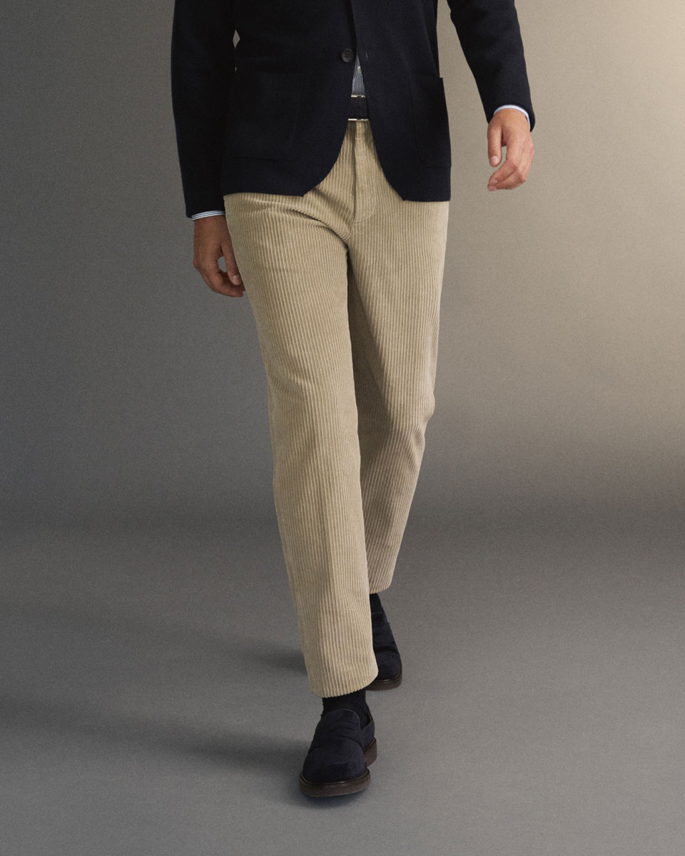 Man wearing beige corduroy pants, navy blue loafers, a dark blazer, and a striped shirt walking against a neutral background.
