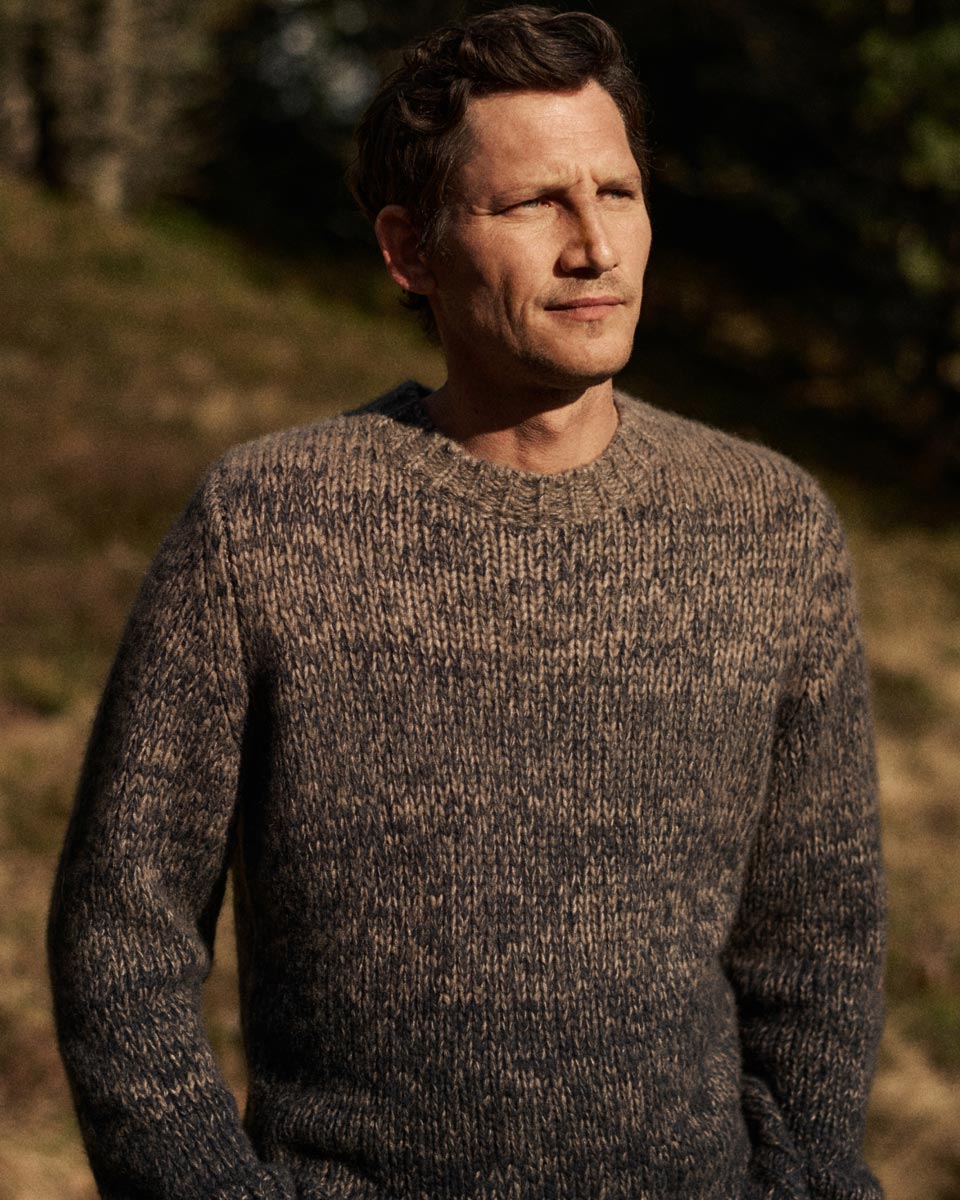 A man with short brown hair wearing a thick, textured brown and gray knit sweater standing outdoors in soft sunlight.