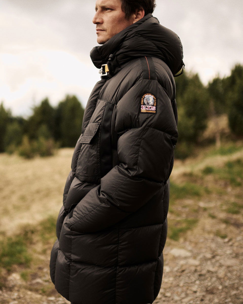Mann in schwarzer, langer Daunenjacke mit Kapuze und Parajumpers-Logo am Arm steht seitlich in natürlicher, bewaldeter Landschaft.