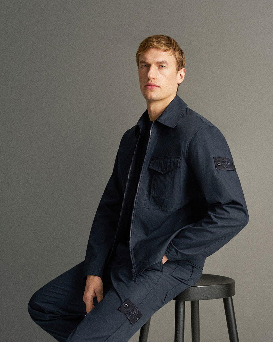 Mann sitzt auf schwarzem Hocker, trägt dunkelblaue Stone Island Jacke und Hose mit Logo-Patches, grauer Hintergrund.