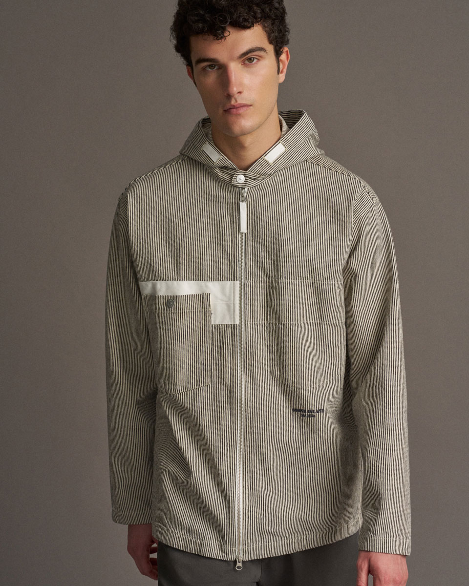 Junger Mann mit kurzen lockigen Haaren trägt beige-weiß gestreifte Jacke mit Kapuze, Reißverschluss und Brusttasche.