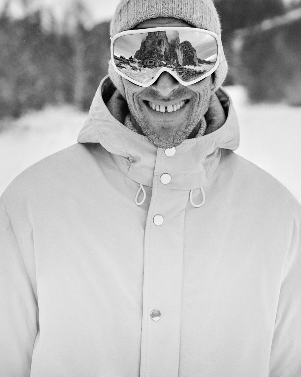 Mann mit Skihelm und Skibrille, die Berge im Spiegel reflektiert, trägt dicke Winterjacke und Mütze, lächelt.