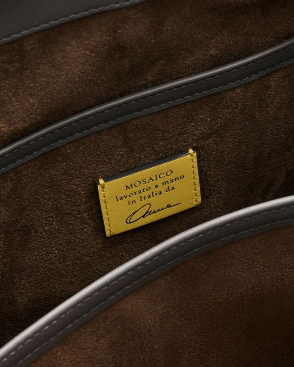 Close-up of a brown suede interior with black leather trim and a yellow label reading 'MOSAICO lavorato a mano in Italia da' with a signature.