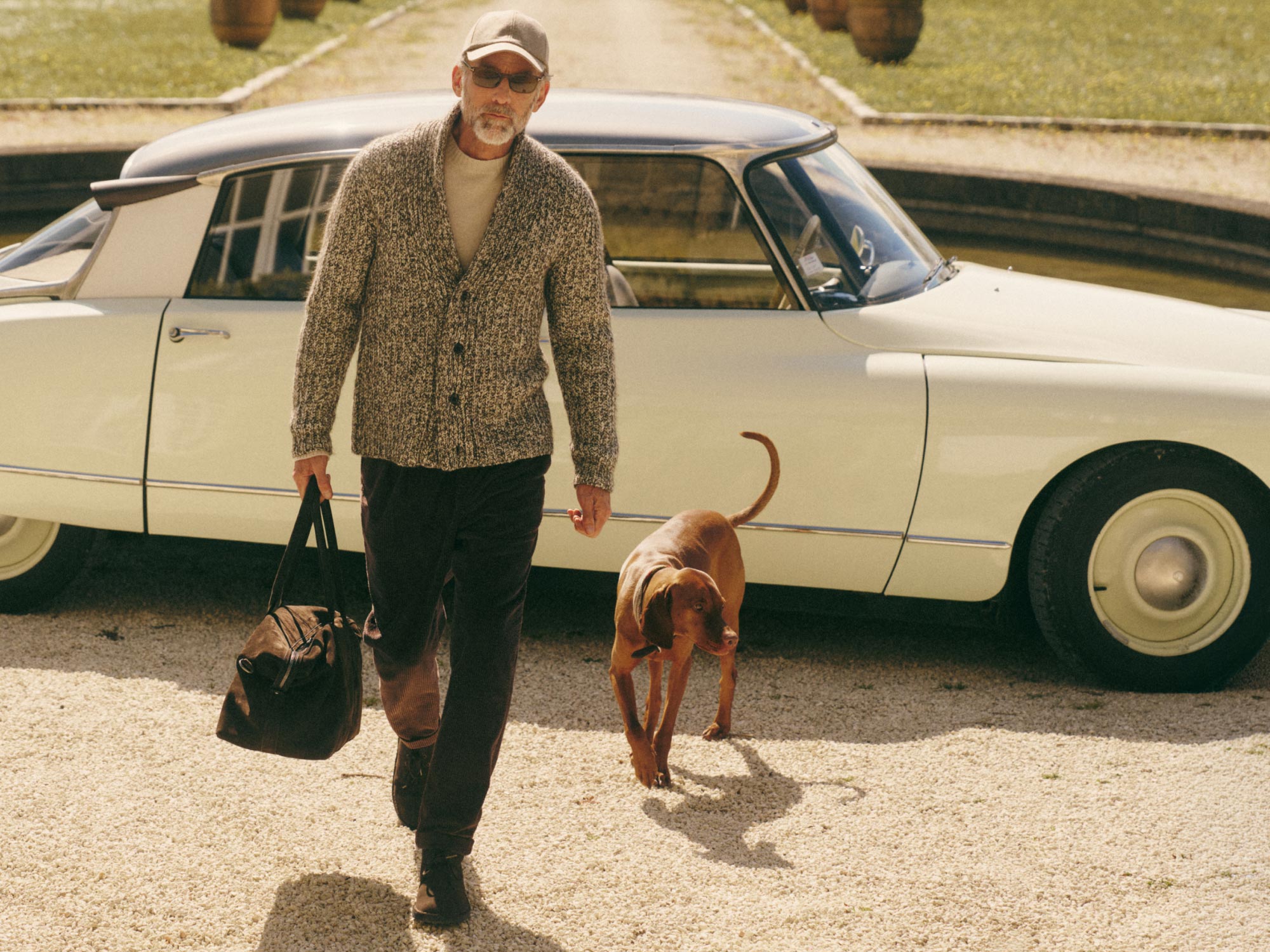 Man in sunglasses and cap wearing a knit cardigan walks a brown dog, carrying a bag, with a vintage white car behind them.