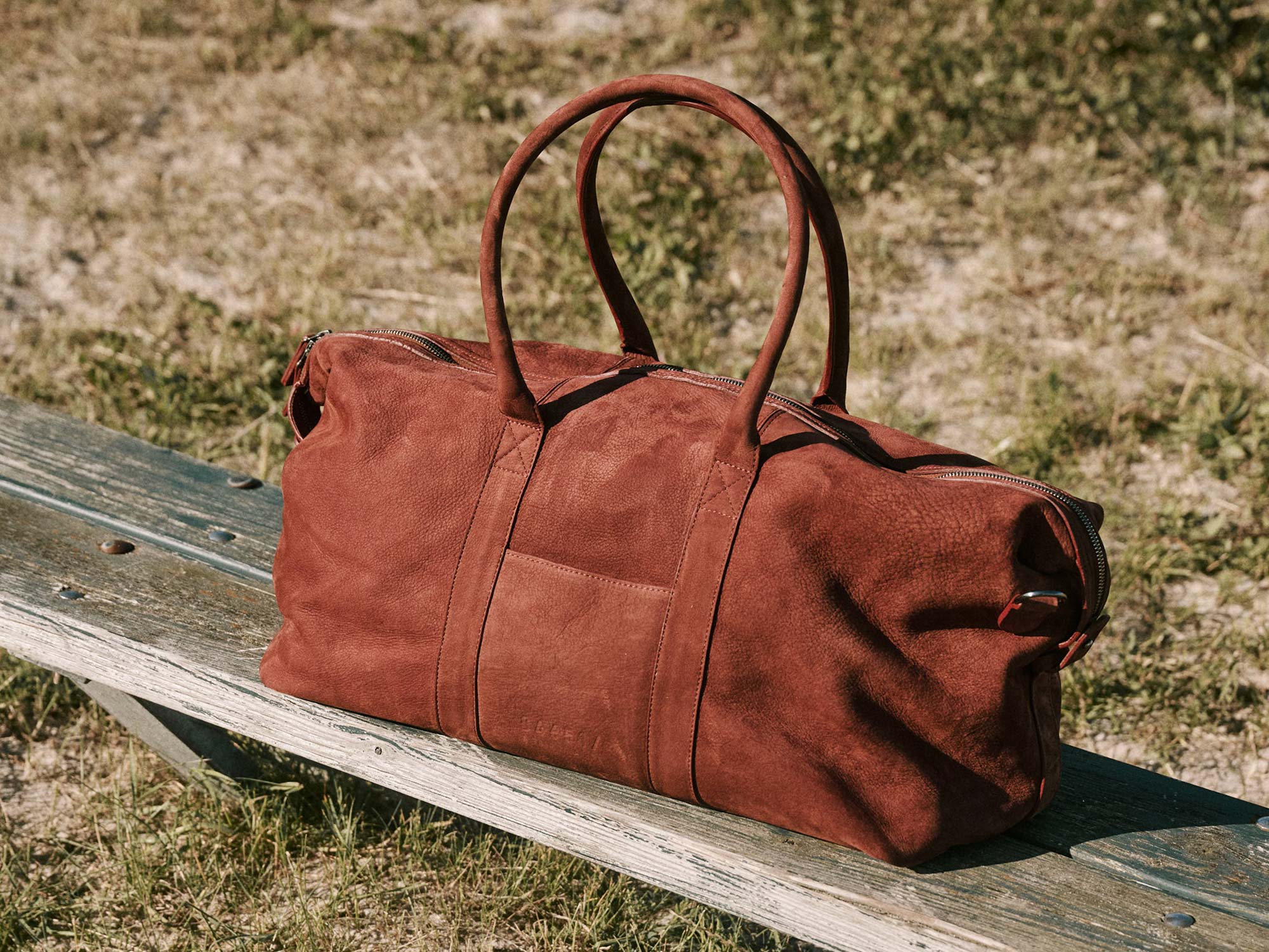 Brown leather duffel bag with dual handles and zipper, resting on a weathered wooden bench outdoors on grass.