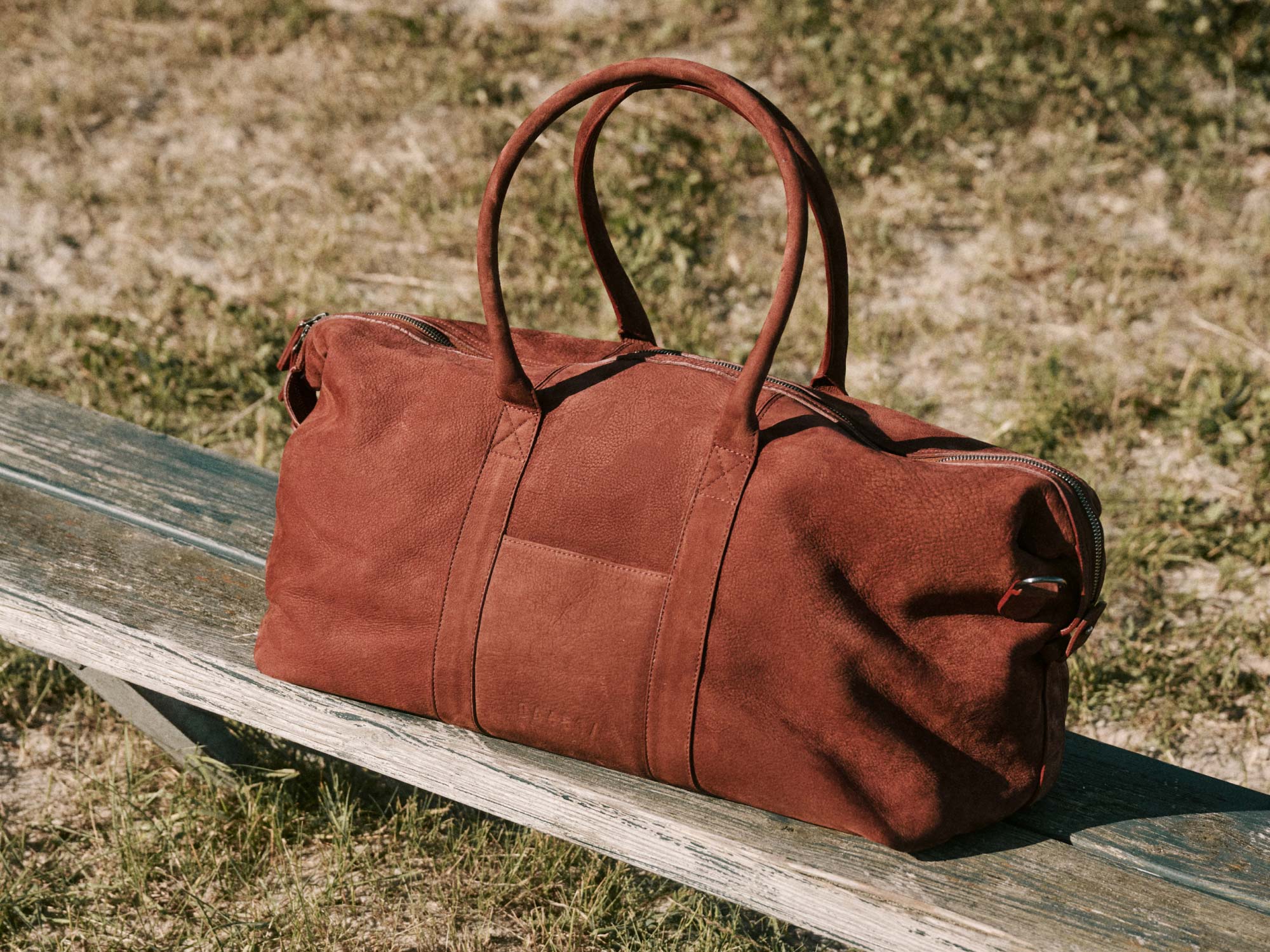 Braune Lederreisetasche mit zwei Henkeln und Reißverschluss liegt auf einer hellen Holzbank im Freien auf Grasboden.