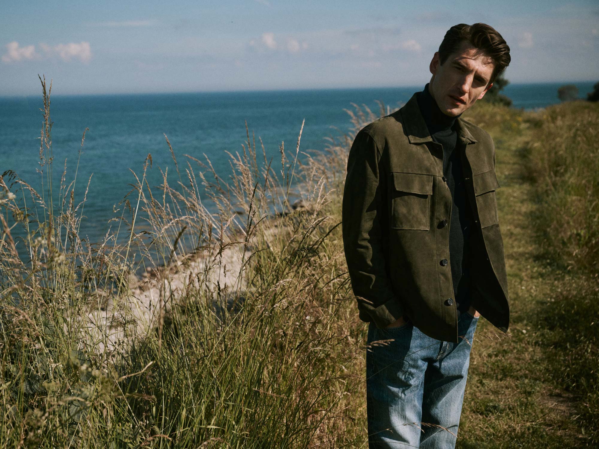 Man in olive green jacket and blue jeans standing among tall grass near a coastline with blue ocean and sky in the background.