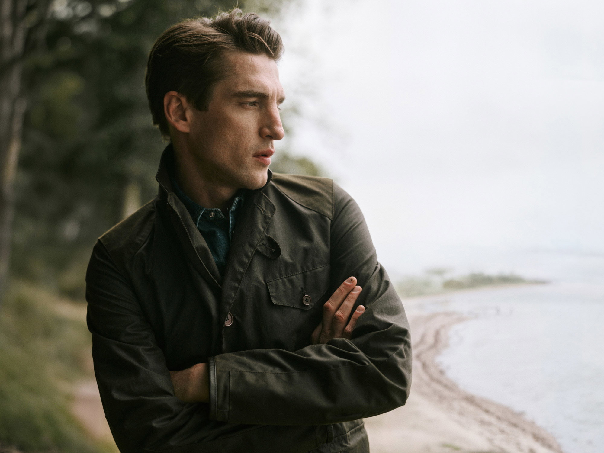 Junger Mann mit dunkelgrüner Barbour-Wachsjacke und blauem Hemd blickt nach rechts, im Hintergrund Wald und Seeufer.