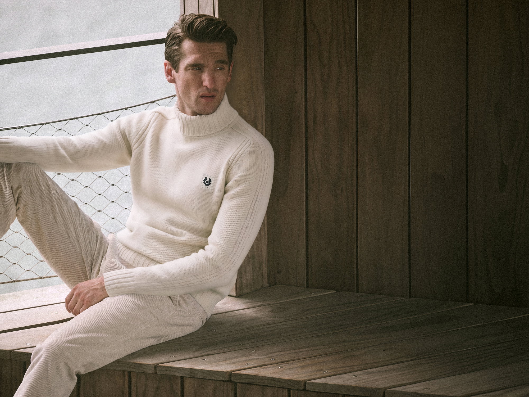Man wearing a white ribbed turtleneck sweater and beige pants sitting on a wooden bench with a netted railing behind.