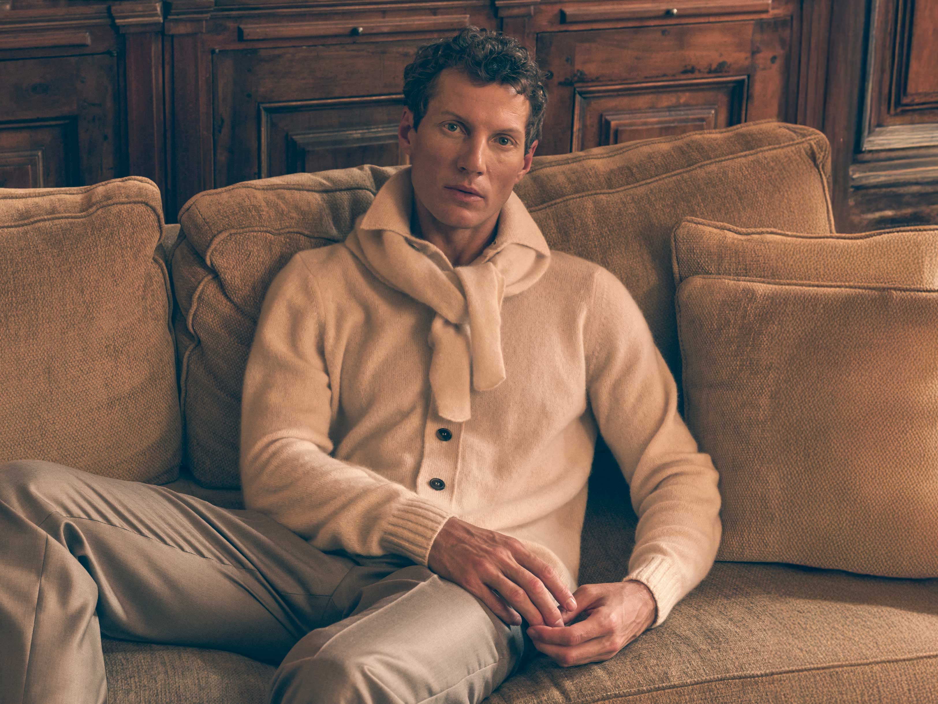 Mann sitzt auf braunem Sofa, trägt beigen Cashmere-Pullover mit Schal, Holzwand im Hintergrund, entspannte Atmosphäre.