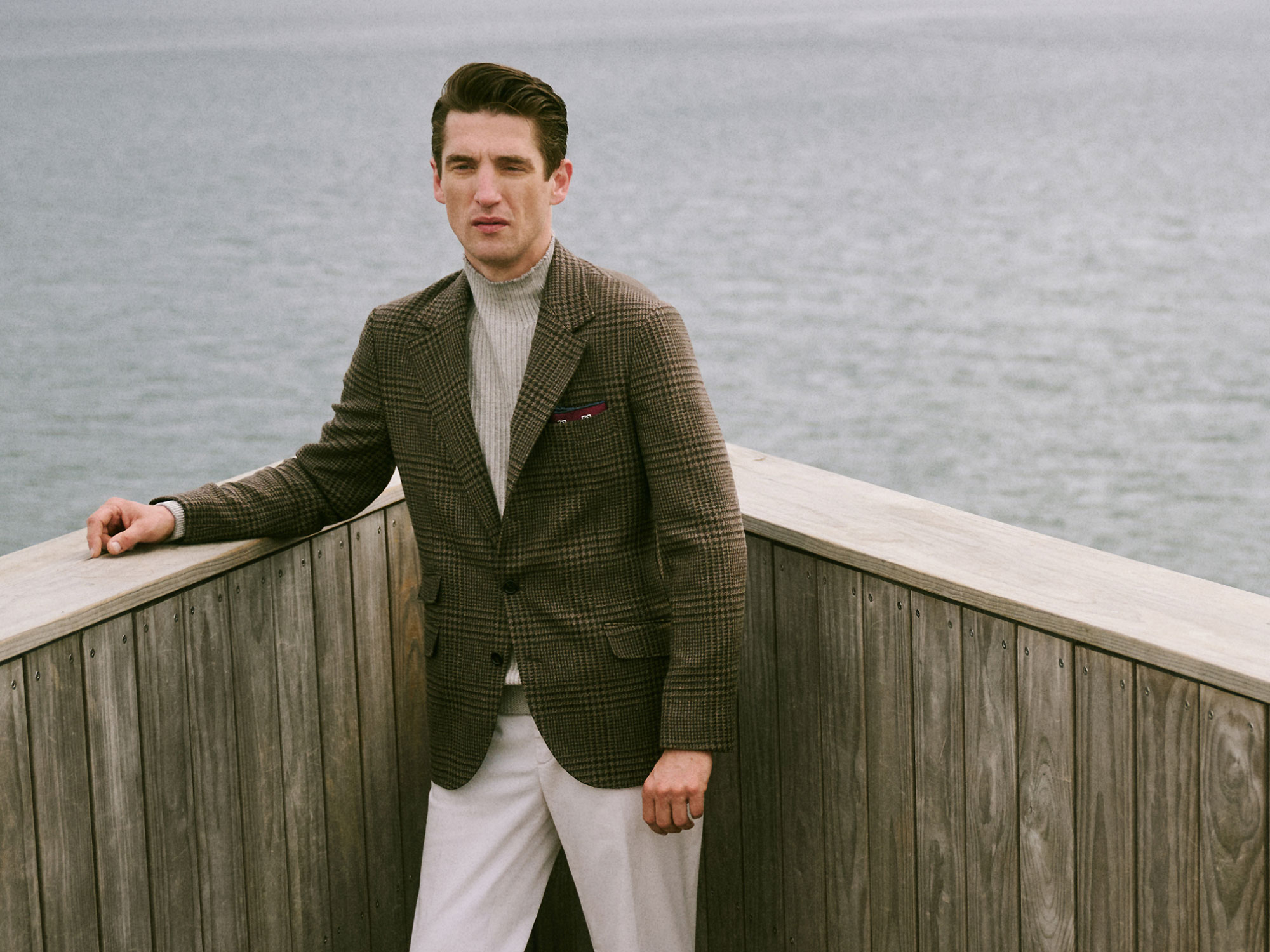 Man wearing a brown checkered blazer, gray turtleneck sweater, and white pants standing on a wooden deck by the water.