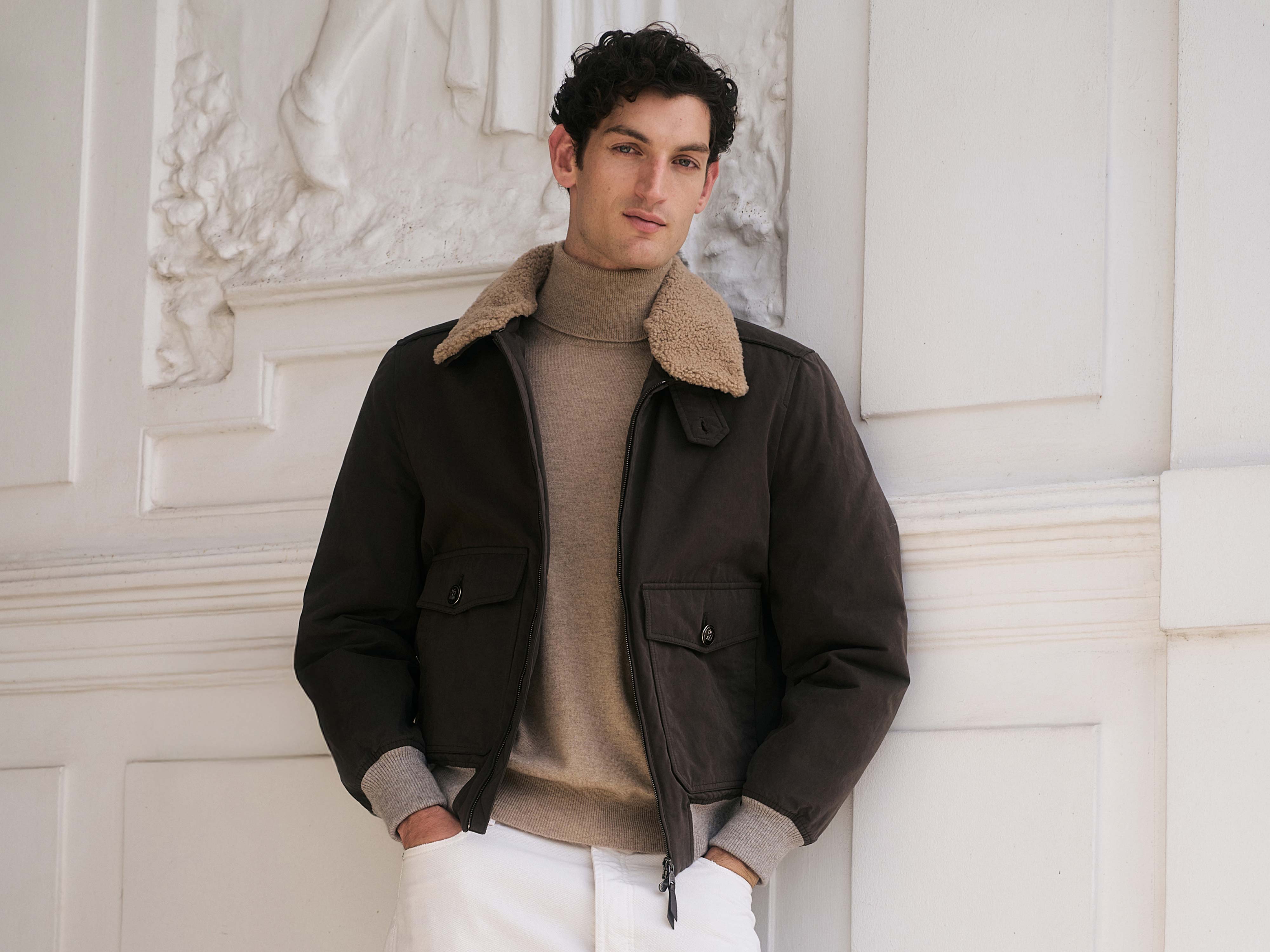 Mann mit dunklem, lockigem Haar trägt braunen Blouson mit Teddyfellkragen, beigen Rollkragenpullover und weiße Hose.