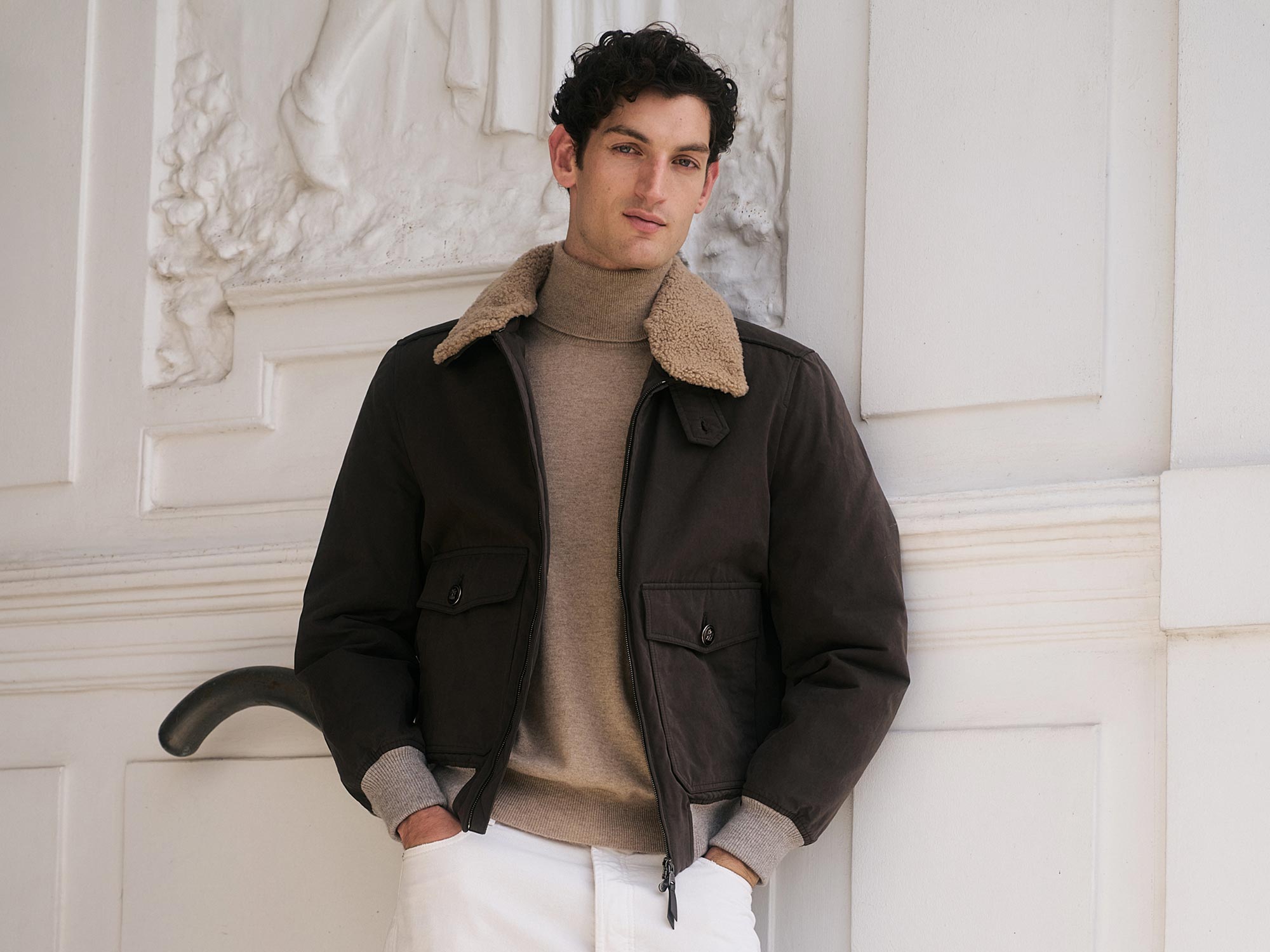 Junger Mann mit dunklem, lockigem Haar trägt braune Jacke mit Teddyfellkragen, beigen Rollkragenpullover und weiße Hose.