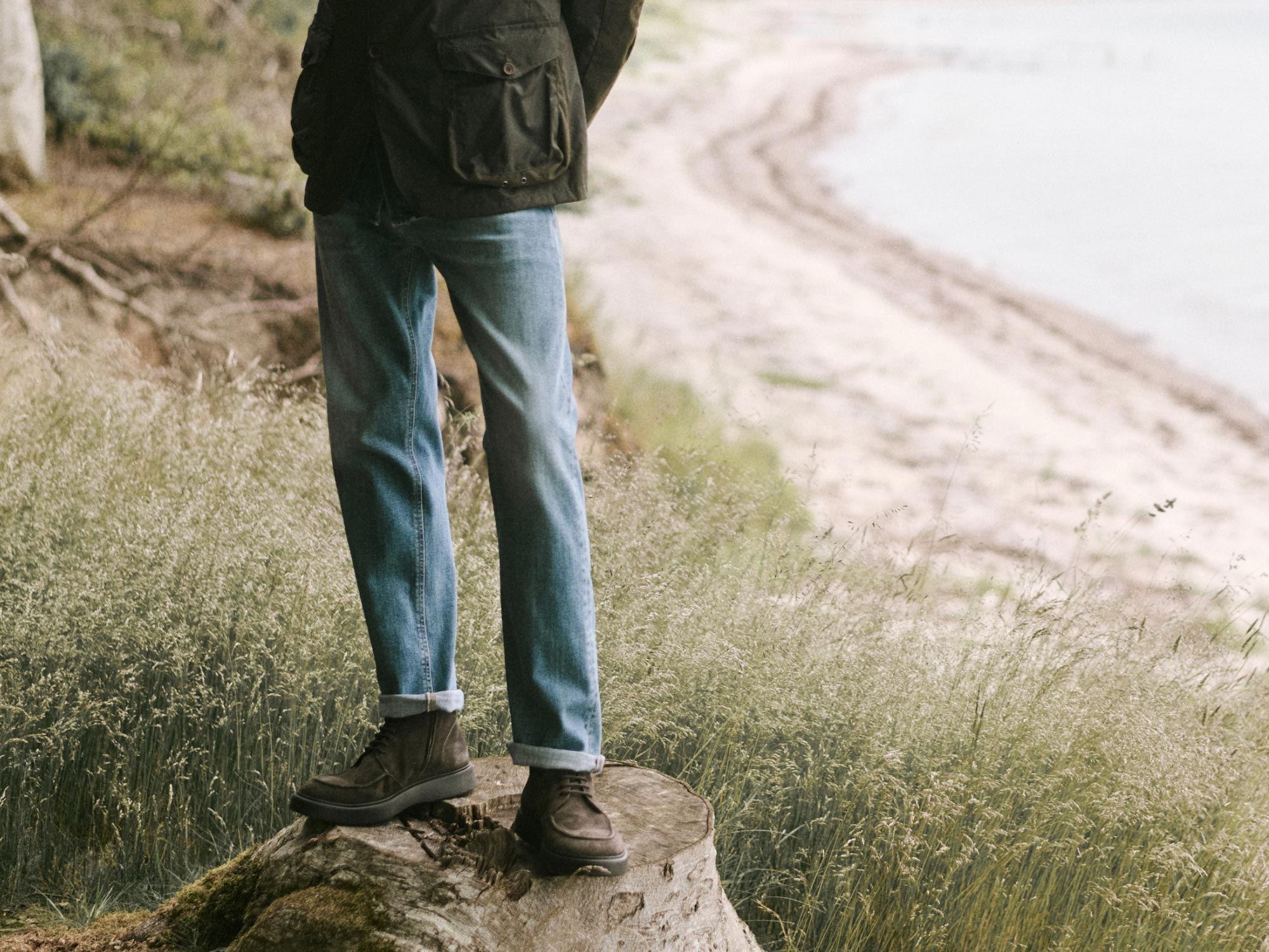 Mann steht auf Baumstumpf am Seeufer, trägt blaue Jeans, braune Boots und grüne Jacke mit Taschen im Wald.
